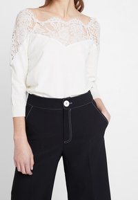 Top bianco con sovrapposizione di pizzo e maniche lunghe, abbinato a pantaloni neri a vita alta con cuciture bianche e chiusura a bottone.
