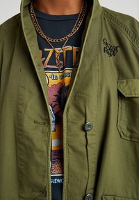 Olive green jacket with a button closure, two pockets, and an embroidered logo. Worn over a black graphic t-shirt featuring band imagery.