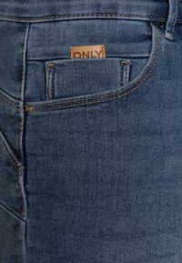 Sininen farkkuhousu, jossa on takatasku, jossa on tanvärinen nahkainen etiketti, jossa lukee "ONLY", kontrastiväriset ompeleet saumoissa ja sileä pinta.