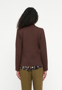Braune, taillierte Jacke mit strukturierter Oberfläche, Kragen und langen Ärmeln. Darunter befindet sich ein gemustertes Oberteil. Olivgrüne Hose rundet den Look ab.