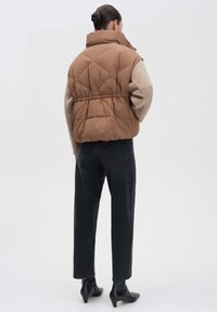 Braune Steppweste mit hohem Kragen, elastischem Bund und Rautensteppung. Kombiniert mit schwarzen geraden Jeans und schwarzen Absatzstiefeln.