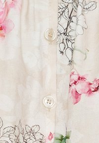 Blomstermønstrede stof i blød beige med pink og grønne accenter. Har tre hvide knapper langs midten og en glat tekstur.