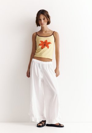 Jeune femme portant un caraco jaune à imprimé fleurs rouges et un pantalon blanc ample avec des ourlets volantés, debout contre un mur blanc.