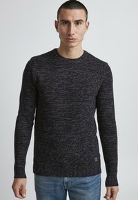 Sötét kötött pulóver, texturált mintával, kerek nyakkivágással, bordás ujjakra és derékra. Világoskék farmernadrággal párosítva.