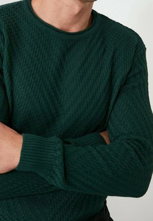 Mężczyzna w ciemnozielonym, teksturowanym, dzianym swetrze z okrągłym dekoltem i ściągaczami na rękawach, z założonymi na piersi rękami na jednolitym tle.