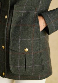 Cappotto in lana a quadri verdi con dettagli in oro, ampie tasche a soffietto e una forma strutturata con cuciture a vista.