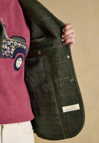 Vihreä tweed-takki, jossa on oranssisävyinen ruutukuvio, tummalla eläinkuosilla vuorattu sisus, ja sisäpuolella etiketti.