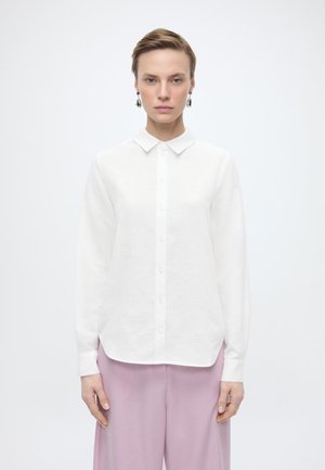 Femeie cu păr scurt, purtând o cămașă albă cu nasturi și pantaloni largi roz deschis, stând în picioare pe un fundal simplu, deschis.