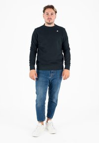 Czarna bluza z okrągłym dekoltem z żebrowanym mankietami, zestawiona z niebieskimi dżinsami i białymi sneakersami. Model pozuje na tle białym.