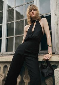 Zwarte jumpsuit met halternek en diepe decolleté, gecombineerd met een kleine zwarte tas. Gouden armbanden accentueren de look.