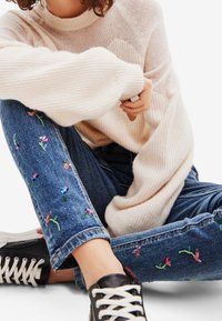 Ljust beige stickad tröja med ribbat mönster, i kombination med blå denimjeans med broderade blommönster och svarta sneakers.