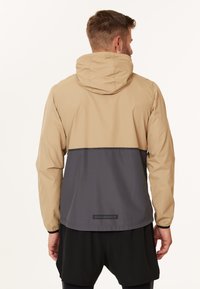 Leichte Jacke mit beigem Oberteil und dunkelgrauem Unterteil. Sie verfügt über eine Kapuze, elastische Bündchen und ein kleines Logo auf der Rückseite.