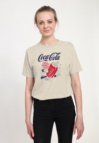 Beżowa koszulka z kolorową grafiką Coca-Cola przedstawiającą puszkę, gwiazdki oraz elementy tekstowe w kolorze czerwonym i niebieskim. Krótkie rękawy i luźny krój.