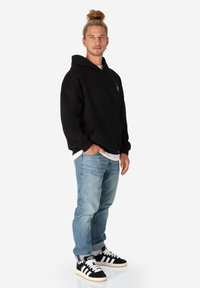 Schwarzer Hoodie mit Kängurutasche, blaue Jeans und schwarze Sneaker mit weißen Streifen. Haare zu einem Dutt frisiert, das Model steht vor einem weißen Hintergrund.