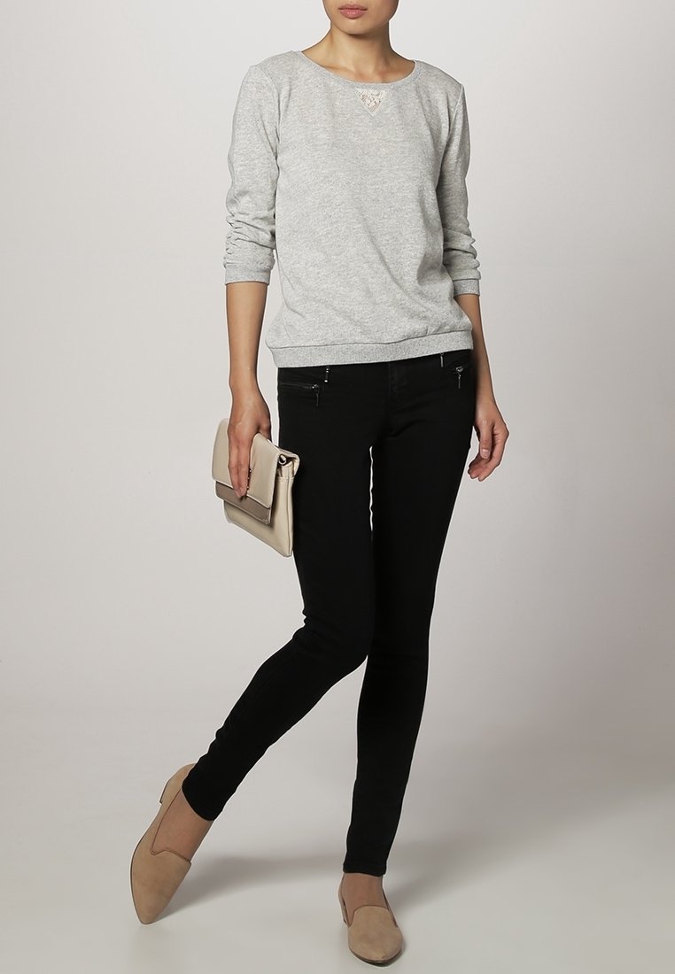 Femme portant un haut gris à manches longues, un pantalon noir moulant, des chaussures beiges, tenant une pochette beige, debout avec une jambe croisée.