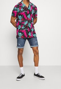 Mies, joka seisoo kädet selän takana, päällä musta, vaaleanpunainen ja sininen kukkakuosiset lyhythihaiset paita, farkkushortsit, valkoiset sukat ja mustat tennarit.