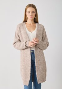 Cardigan en tricot marron clair avec une texture douce, à l'avant ouvert et à manches longues, porté par-dessus un t-shirt blanc à col en V et un jean bleu.