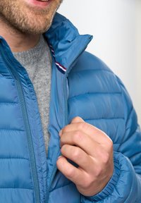 Homme portant un pull gris et une doudoune bleue qui ferme la fermeture éclair de la veste, gros plan sur la main et le col de la veste.