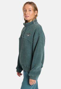 Sudadera de media cremallera en color verde azulado con textura acanalada, puños y cintura elásticos. Presenta un logo bordado en el pecho. Tejido suave.