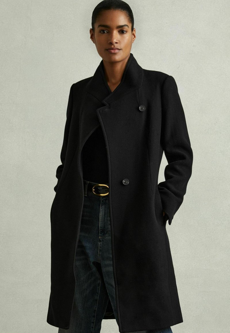 Cappotto in lana nera con design doppiopetto, ampi rever e una sottile trama. Abbinato a jeans scuri a vita alta e una cintura dorata.