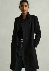 Abrigo de lana negro con cuello de solapa, doble botón y bolsillos laterales. Combinado con jeans oscuros y una blusa negra, que resalta una silueta elegante.