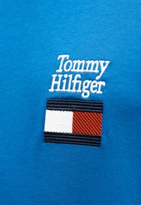 Μπλε βαμβακερό μπλουζάκι με λευκό κεντημένο κείμενο "Tommy Hilfiger" και ορθογώνιο λογότυπο με κόκκινες, λευκές και ναυτικές ρίγες.