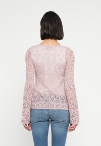 Ανοιχτό ροζ πουλόβερ από δαντέλα με μακριά μανίκια, στρογγυλή λαιμόκοψη και περίπλοκα διαμάντια μοτίβα. Φορεμένο πάνω από στενά μπλε τζιν.