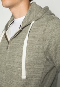Grijze zip-up hoodie van een gestructureerde stof, met witte trekkoorden en een contrasterende binnenvoering bij de kraag.