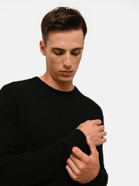Maglione nero a maglia con scollatura a girocollo, maniche lunghe e polsini a coste. Texture liscia, design aderente, con le mani incrociate vicino al petto.