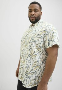 Camisa de manga corta en tejido ligero con una base crema y un patrón de hojas en amarillo y azul, que presenta un cuello abotonado y un corte relajado.