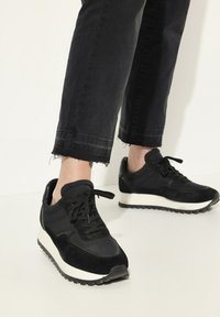 Sneakers neri con un mix di camoscio e materiale sintetico, caratterizzati da una suola intermedia bianca, punta rotonda e design con lacci. Indossati con jeans neri a vita alta.