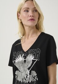 Melna kokvilnas T-kreklu ar V veida kakla izgriezumu, ar sudrabainu grafikas dizainu, uz kura attēlota ērglis un rozes, virs kura rakstīts "MŪŽĪGI SPĒCĪGS".