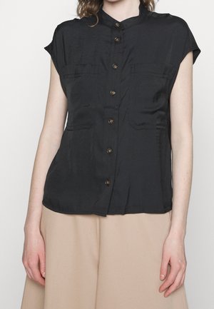 Vrouw die een zwarte blouse met korte mouwen, een mandariinkraag en een beige rok draagt, staand tegen een effen achtergrond.