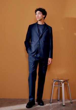 Junger Mann in einem dunkelblauen Anzug und schwarzen Schuhen steht neben einem silbernen Metallhocker vor einer schlichten braunen Wand.