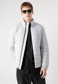 Gai pelēks, quilted pūkains mētis ar augstu apkakli un elastīgiem aprocēm, uzvilkts virs melna pogājama krekla; tam ir priekšējais zip un logo detaļa.