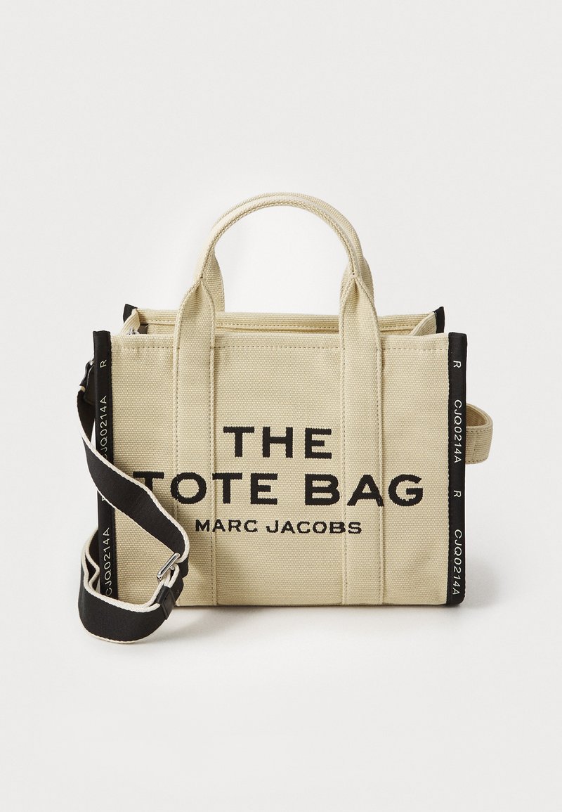 Beżowa torba płócienna z czarnym regulowanym paskiem na ramię, krótkimi uchwytami oraz napisem "THE TOTE BAG MARC JACOBS" na froncie.
