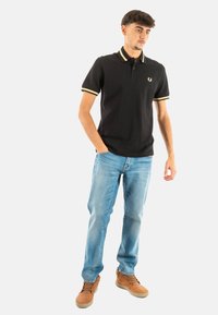 Polo nero con accenti gialli, maniche corte e una texture morbida. Abbinato a jeans azzurri chiari e scarpe marroni, in piedi in modo informale.