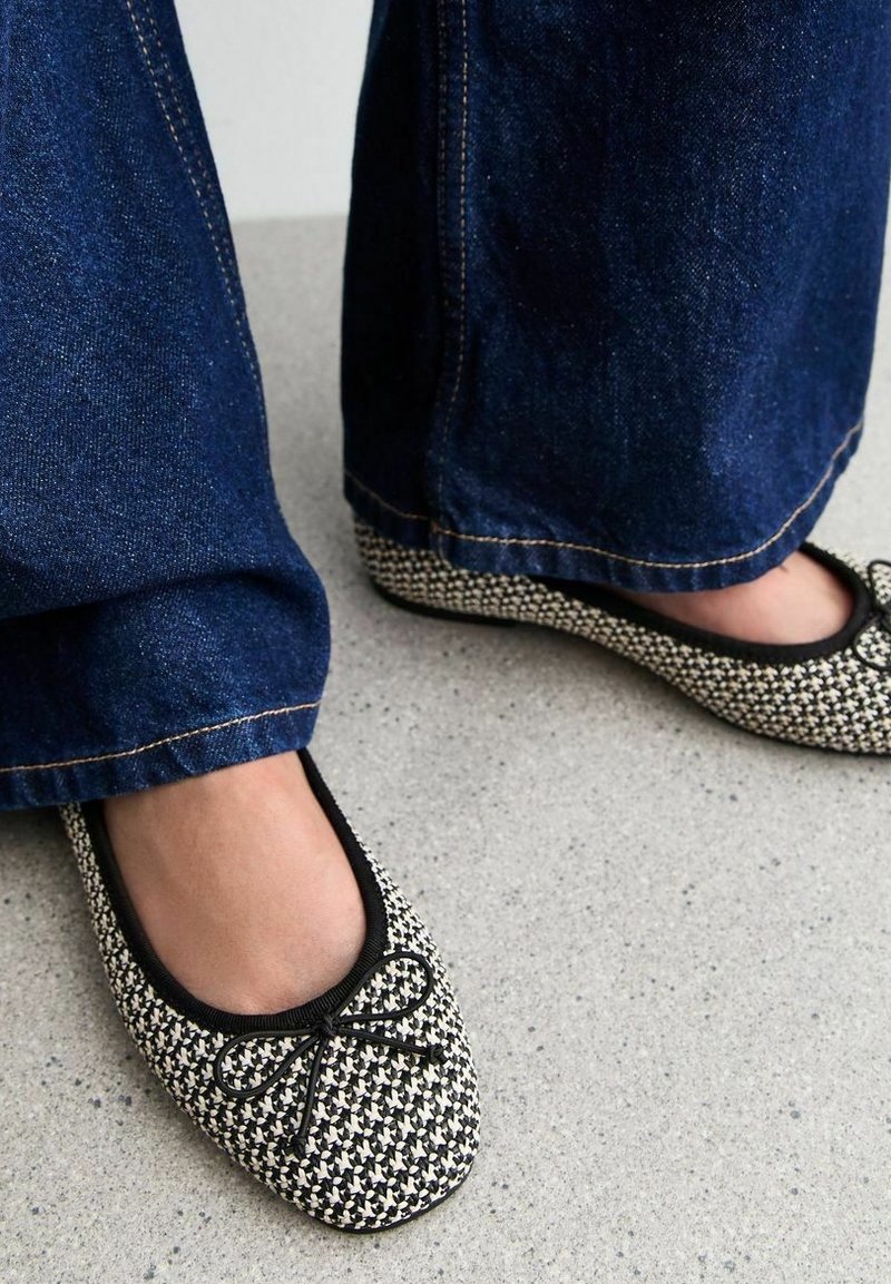 Pieds portant des ballerines à motifs noir et blanc avec bordure noire et nœuds, assortis à un jean bleu foncé à jambes larges sur un sol gris.