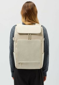 Beige ryggsäck med lockstängning, justerbara remmar, en framficka och en slät yta, visad mot en neutral bakgrund.