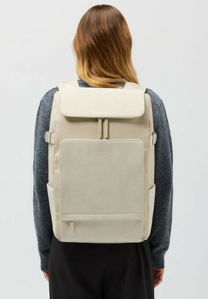 Beige reppu, jossa on läppä suljenta, säädettävät hihnat, etutasku ja sileä pinta, esiteltynä neutraalia taustaa vasten.