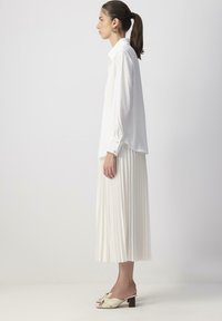 Femeie cu păr brunet prins în coadă de cal, îmbrăcată cu o cămașă albă cu mânecă lungă, fustă midi plisată albă, cercei aurii și sandale cu toc crem, stând de profil.