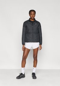 Μαύρο μπουφάν με επένδυση, φερμουάρ μπροστά, ψηλό γιακά, ελαστικές μανσέτες και λογότυπο Nike. Φορεμένο πάνω από μαύρο τοπ και λευκό σορτς, συνδυασμένο με μαύρα αθλητικά παπούτσια.