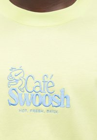 Svetložltá bavlnená mikina s modrým vyšívaným logom „Café Swoosh“ a textom „HORÚCE. ČERSTVÉ. DENNE.“ pod ním. Okrúhly výstrih.