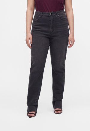 Femme portant un jean noir taille haute, un haut bordeaux et des talons ouverts noirs, debout les mains relâchées le long du corps sur fond blanc.