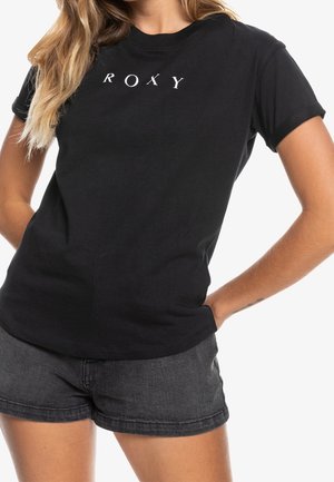 Femme portant un t-shirt ROXY noir associé à un short en denim gris foncé, mains posées sur les hanches, sur fond blanc.