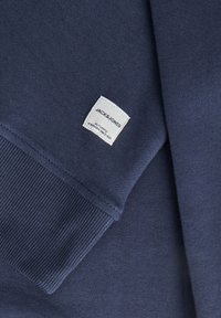 Mörkblå sweatshirt med ribbade ärmslut och nederkant. Har en vävd etikett sydd i den nedre delen, som visar varumärket och autenticitetens detaljer.