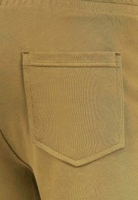 Pantaloni khaki con un tessuto texturizzato, una tasca posteriore rettangolare con bordi angolari e dettagli di cucitura visibili, che mettono in risalto un colore uniforme.