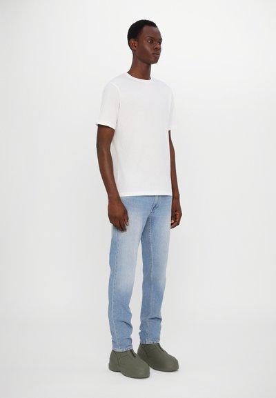 Weißes T-Shirt mit kurzen Ärmeln, hellblaue Jeans und grüne Slip-on-Schuhe. Einfaches Design, glatte Textur und entspannte Passform.