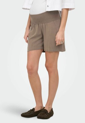 Naine kannab pruune elastse vöökohaga lühikesi pükse, valget toppi, valget jopet ja tumedat pruunid mokassiinsaapaid, seisab ühevärvilise tausta ees.