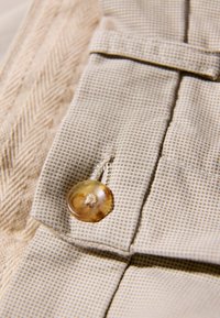 Tissu beige texturé avec un motif en grille, présentant un bouton rond de couleur ambre cousu sur le bord d'un rabat.